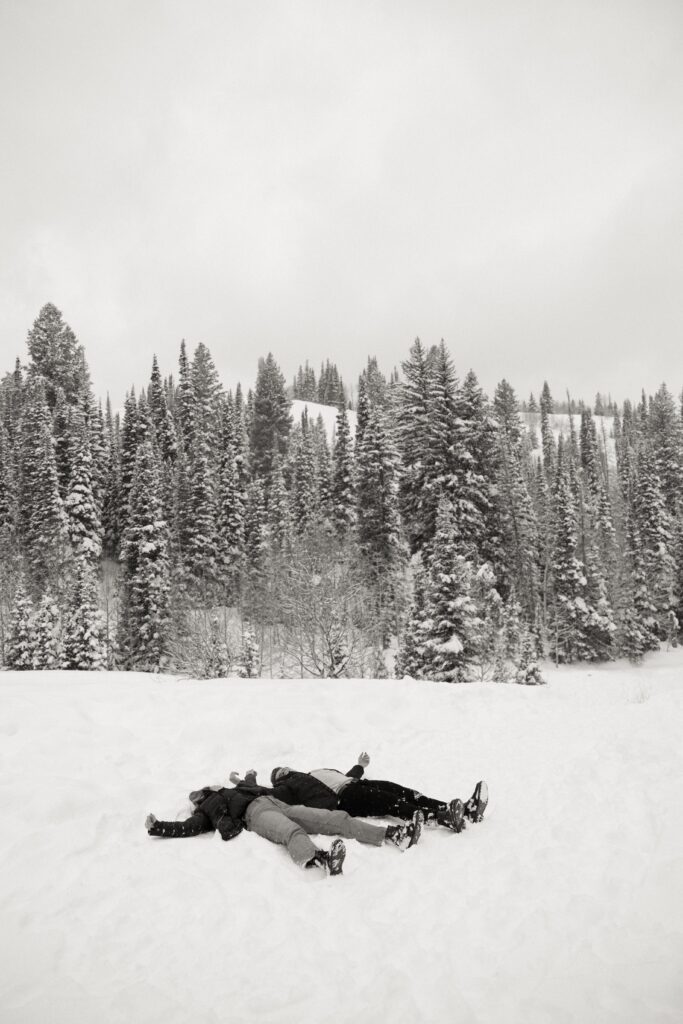 A couple making snow angles with the snow covered trees in the background.