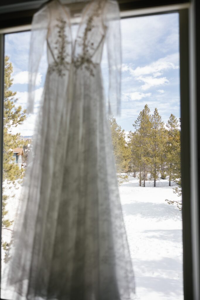 A wedding dress hung in the window, the snow and pine trees of Colorado can be seen through the window.